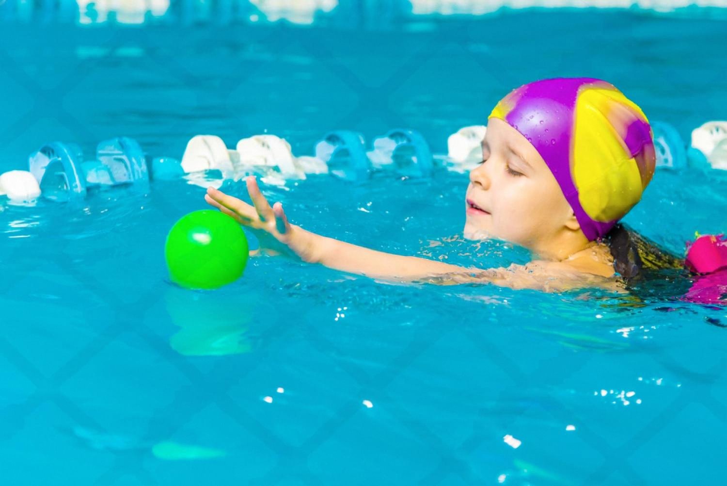 Young child in a swim cap reaching for a ball during a swimming lesson